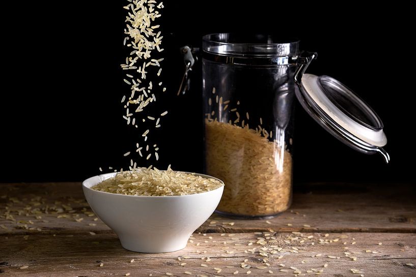 rice grains falling into a white bowl beside a glass jar with rice on a rustic wooden table against  by Maren Winter