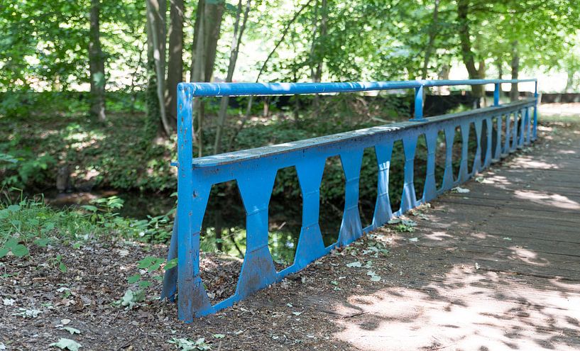 old blue metal bridge with wooden planks over a small river in a park with lots of greenery, trees a by ChrisWillemsen