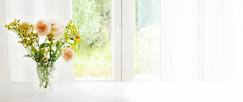 Bouquet from orange roses and rue in glass vase at the window in front of a white curtain, panoramic