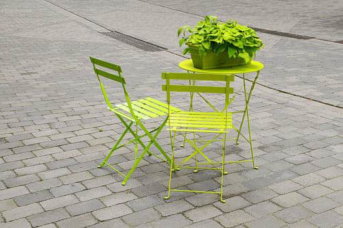 hellgrüne Gartenstühle und ein Klapptisch mit einem grünen Blumenkasten auf dem grau gepflasterten M
