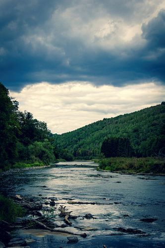 A river on a dark cloudy day in the Belgian Ardennes surrounded by mountains and forest.