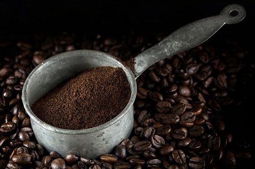 small metal pot with ground coffee on a heap of whole roasted coffee beans, dark background, closeup