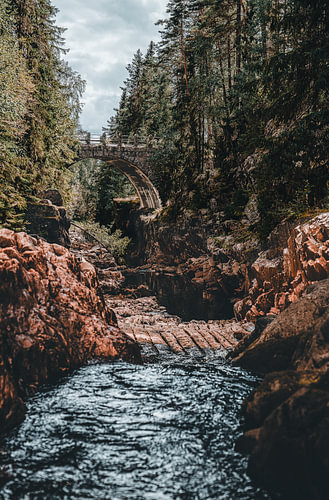 Photographie verticale au style « moody » représentant le pont en arc historique en pierre et le flottage du bois dans les gorges de la rivière Brattfallet, dans le Värmland, en Suède