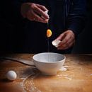 older women hands breaking an egg with falling egg yolk over a bowl, wooden kitchen board, dark back by Maren Winter thumbnail