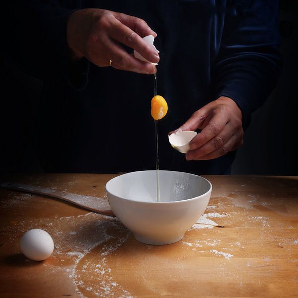 older women hands breaking an egg with falling egg yolk over a bowl, wooden kitchen board, dark back by Maren Winter