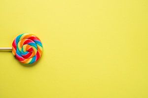 Regenbogenfarben auf einem Lutscher vor einem leuchtend gelben Hintergrund – fröhliche Kinder- und Food-Fotografie