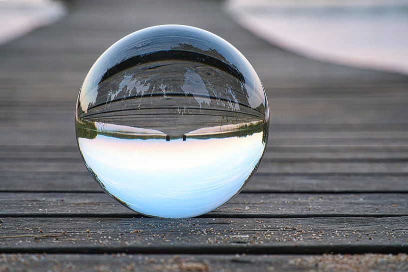 Glass ball on a footbridge reaching into a lake by Martin Köbsch