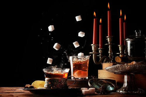 in Teetassen aus Glas fallende Zuckerwürfel auf einem rustikal gedeckten Tisch mit Kerzen und Bonbon