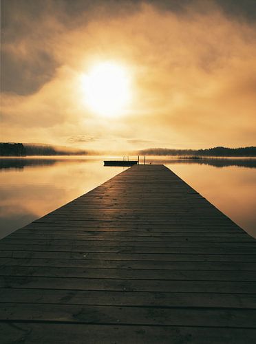 A wooden jetty on the lake in the morning mist, flooded with sunlight and a calm reflection.