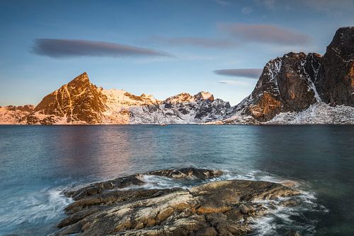 Eisplatten auf dunklem Fels mit See und schneebedeckten steilen Bergen im Reinefjord auf den Lofoten