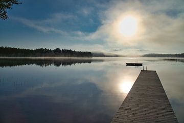 A wooden jetty on the lake in the morning mist, flooded with sunlight and a calm reflection. by Martin Köbsch