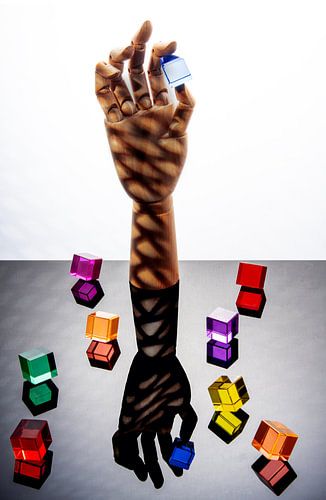 Hölzerne Hand, die einen blauen Würfel zwischen Daumen und Zeigefinger hält, umgeben von 8 mehrfarbigen Würfeln. von Gerhard de Wit