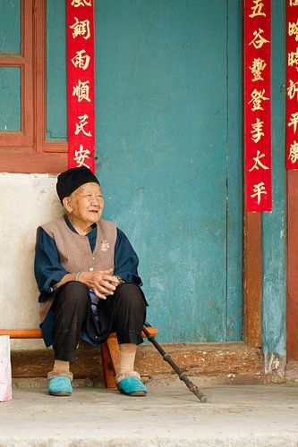 Old Chinese woman sits quietly on a bench in front of a turquoise wall