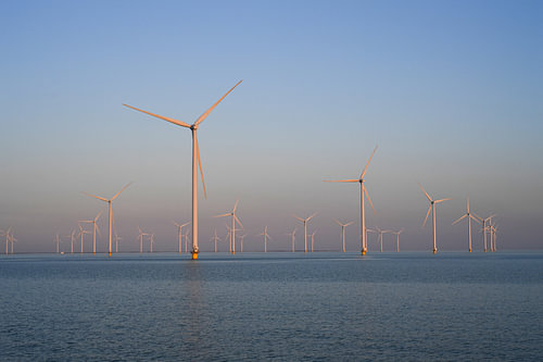 Fryslan Wind Farm: a powerful wind farm rises serenely from the calm water’s surface, bathed in the warm, golden light of the setting sun. The sleek silhouettes of the wind turbines form a stunning contrast with the soft pastel hues of the sky. A vision of perfect harmony between human innovation and the timeless beauty of nature.