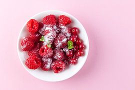 Himbeeren und rote Beeren in einer weißen Schale vor einem rosa Hintergrund.