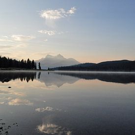 A serene lake in Canada, surrounded by majestic mountains and lush green trees, under a blue sky during sunrise, with dew on the surface of the water and one boathouse in the foreground. The calm water reflects the landscape, creating a sense of symmetry and balance. The landscape exudes peace and serenity. by UMA Digital NL
