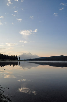 Un lac serein au Canada, entouré de montagnes majestueuses et d'arbres verdoyants, sous un ciel bleu au lever du soleil, avec de la rosée à la surface de l'eau et un hangar à bateaux au premier plan. L'eau calme reflète le paysage, créant un sentiment de symétrie et d'équilibre. Le paysage respire la paix et la sérénité.