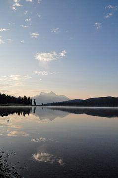 A serene lake in Canada, surrounded by majestic mountains and lush green trees, under a blue sky during sunrise, with dew on the surface of the water and one boathouse in the foreground. The calm water reflects the landscape, creating a sense of symmetry and balance. The landscape exudes peace and serenity. by UMA Digital NL