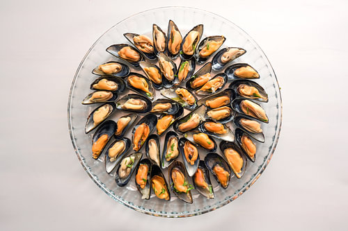 Mit Zwiebeln und Kräutern gekochte Muscheln, serviert auf einer runden Glasplatte, grauer Hintergrun