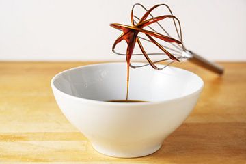 Brown sugar syrup flows from a wire whisk into a white bowl, baking at home concept, wooden table an by Maren Winter
