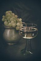 A glass of white wine in a Roman glass with a metal pitcher with light grapes in the background