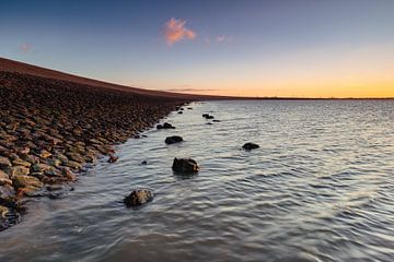 De stenen dijk aan de Waddenzee in Noord-Holland wordt mooi verlicht door de ondergaande zon. van Bram Lubbers