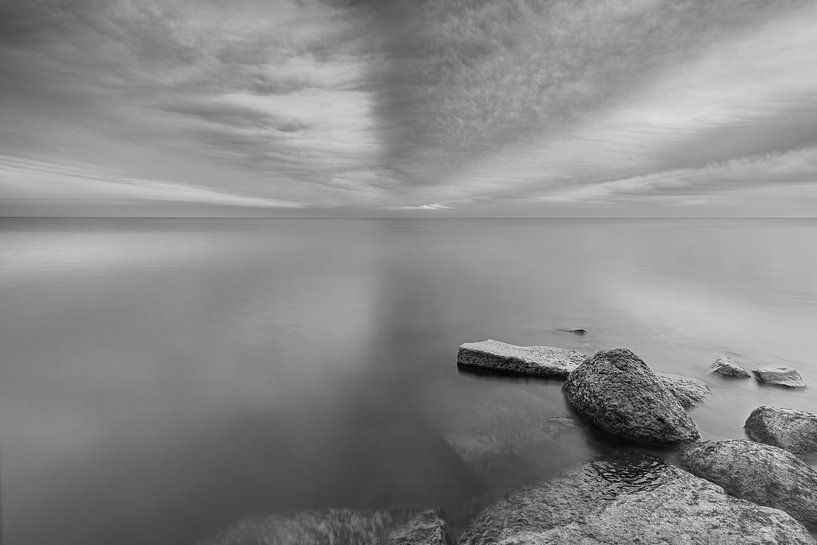 Zwartwit foto van dreigende gestreepte wolken boven een rustig Markermeer met stenen als voorgrond van Bram Lubbers