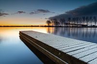 Snow-covered jetty and row of trees reflect in the calm lake during sunrise