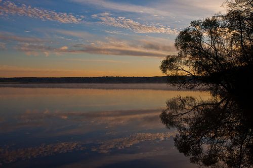 Dawn over the quiet water surface of the lake. The morning blue sky is lit by the orange light of th