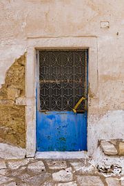 Sandfarbene Steinmauer mit einer blauen Metalltür und Eisenverzierungen im Fenster