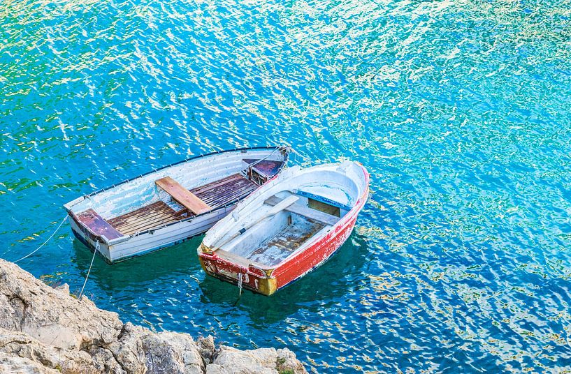 Twee oude roeiboten aangemeerd aan de zeekust met turquoise gekleurd water van Alex Winter