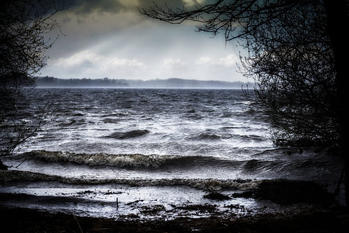 See mit dunklen Wellen am Ufer bei schlechtem Sturmwetter im Wald und Sonnenstrahlen am Himmel, Land