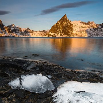 Ijsplaten op donkere rots met meer en met sneeuw bedekte steile bergen in de Reinefjord op de Lofote van Robert Ruidl