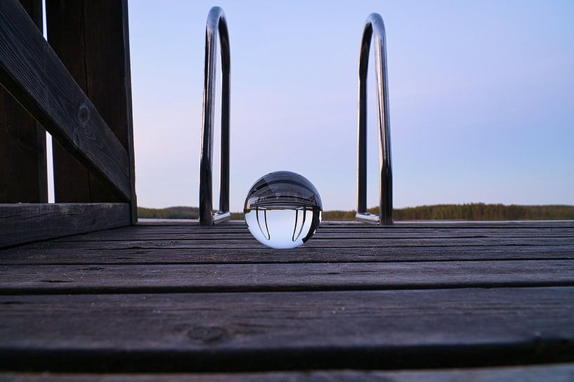 Glass ball on a footbridge reaching into a lake by Martin Köbsch