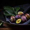 Vers geoogste pruimen met takje en blaadjes in een zwart schaaltje met van Maren Winter