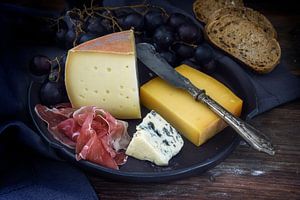 Eetbordje met diverse kaassoorten, luchtgedroogde ham, blauwe druiven en brood op donker rustiek hou