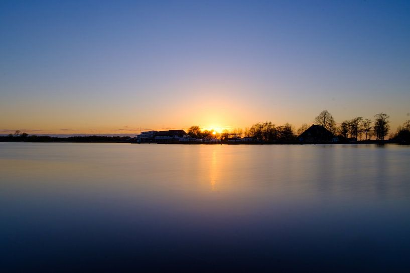 Untergehende Sonne an einem See, Blick auf Boote und Hütten. von Lidewij Olive