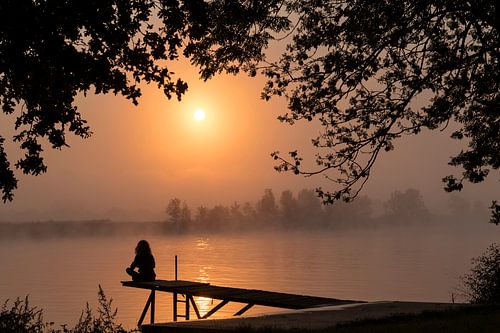 silhouette girl at wooden jetty during sunset in the early morning over the river maas in limburg in