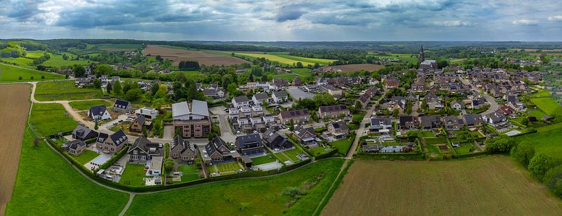 Panoramisch uitzicht op een vredig Limburgs dorp te midden van glooiende heuvels, groene velden en karakteristieke woningen onder een dramatisch wolkendek in de lente. van byphotographyms