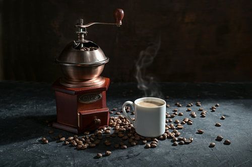 Manual wooden coffee grinder, white cup with the steaming hot drink and some roasted beans on a dark