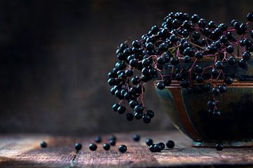 Black elderberries bunch (Sambucus nigra) in an old clay bowl and some berries on a rustic wooden ta by Maren Winter