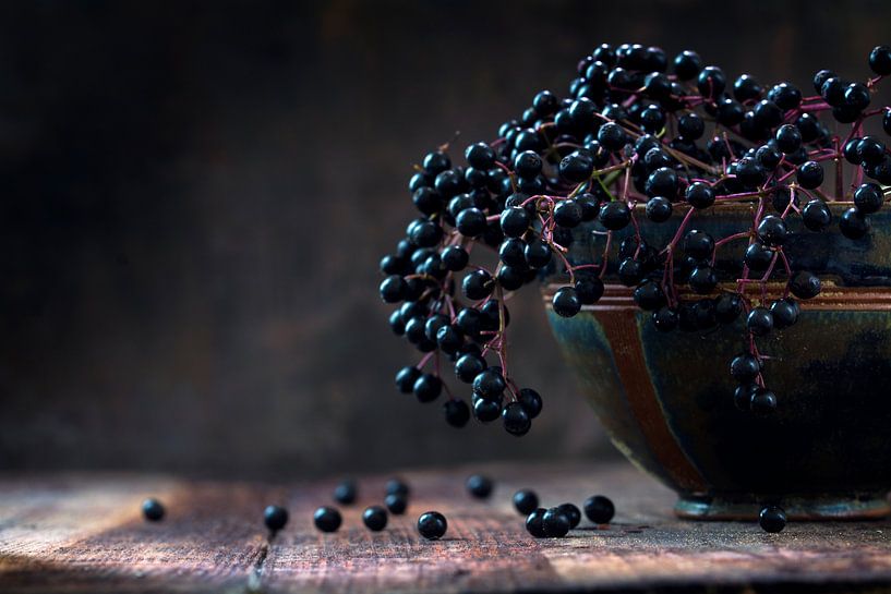 Black elderberries bunch (Sambucus nigra) in an old clay bowl and some berries on a rustic wooden ta by Maren Winter