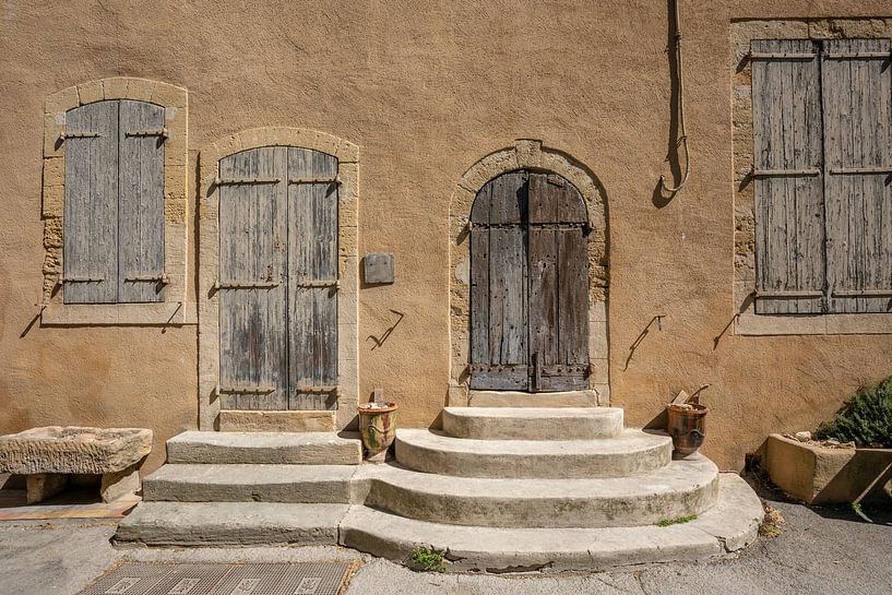 Yellow wall with two doors with pavement and two and windows in village of Loumarin, France by Joost Adriaanse