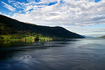 Nordfjord in West-Norwegen mit glattem Wasser und hochaufragenden Gipfeln, an deren Fuß einige Bauernhöfe im Sonnenschein erstrahlen