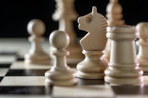 White wooden chess pieces with focus on the knight on a chessboard against a black background, copy 
