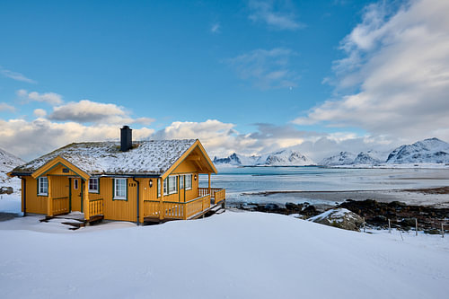 hoelzernes gelbes Haus oder Huette in Winterlandschaft bei stürmischen Wetter  mit Meer und Bergen