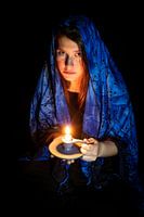 Sad young woman with candle and blue headscarf against black background