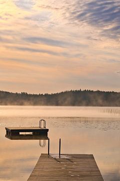 A wooden jetty on the lake in the morning mist, flooded with sunlight and a calm reflection. by Martin Köbsch