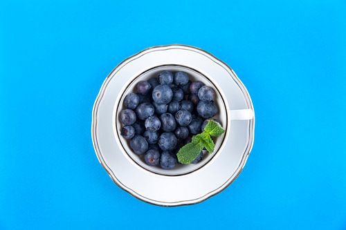 Blauwe bessen in een witte kop op schotel tegen blauwe achtergrond.
