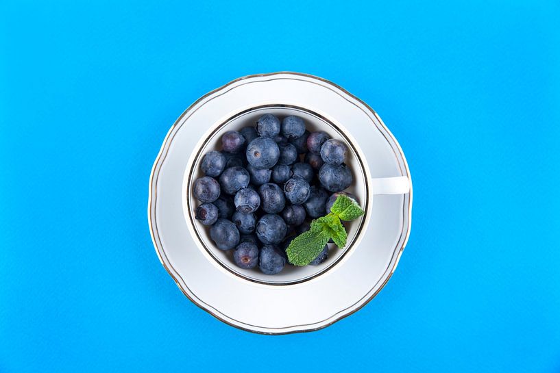 Blueberries in a white cup on saucer against a blue background. by Ans van Heck